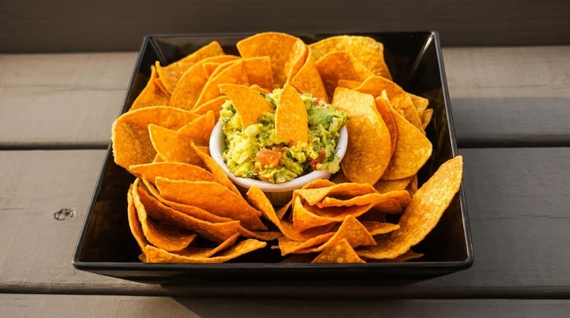 Guacamole dip with tortilla chips in a black square dish on a wooden table.