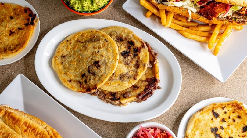 Assortment of Salvadoran dishes including pupusas, a sandwich with fries, guacamole, and curtido.
