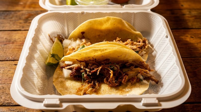 Two street tacos with shredded meat, onions, and lime in a white to-go container.