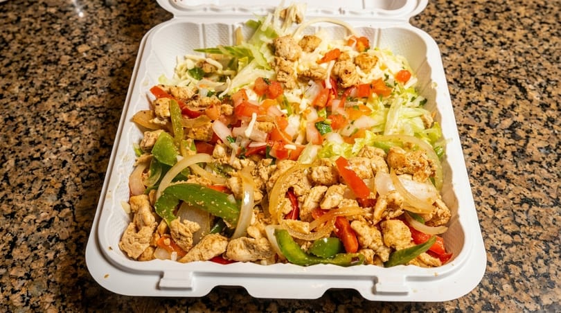 Chicken fajita bowl with peppers, onions, lettuce, and pico de gallo in a white takeout container.