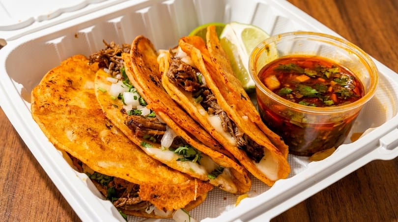 Four cheesy birria tacos with consommé and lime in a white takeout container.