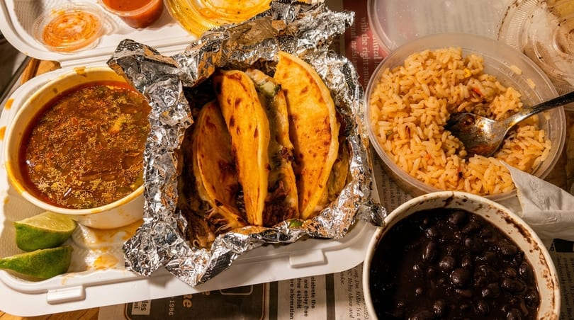 Mexican takeout meal featuring tacos, birria soup, rice, black beans, and lime wedges.
